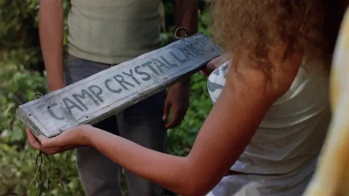 A camp counsellor holds awooden Camp Crystal Lake sign in a screenshot from the original Friday the 13th (1980).